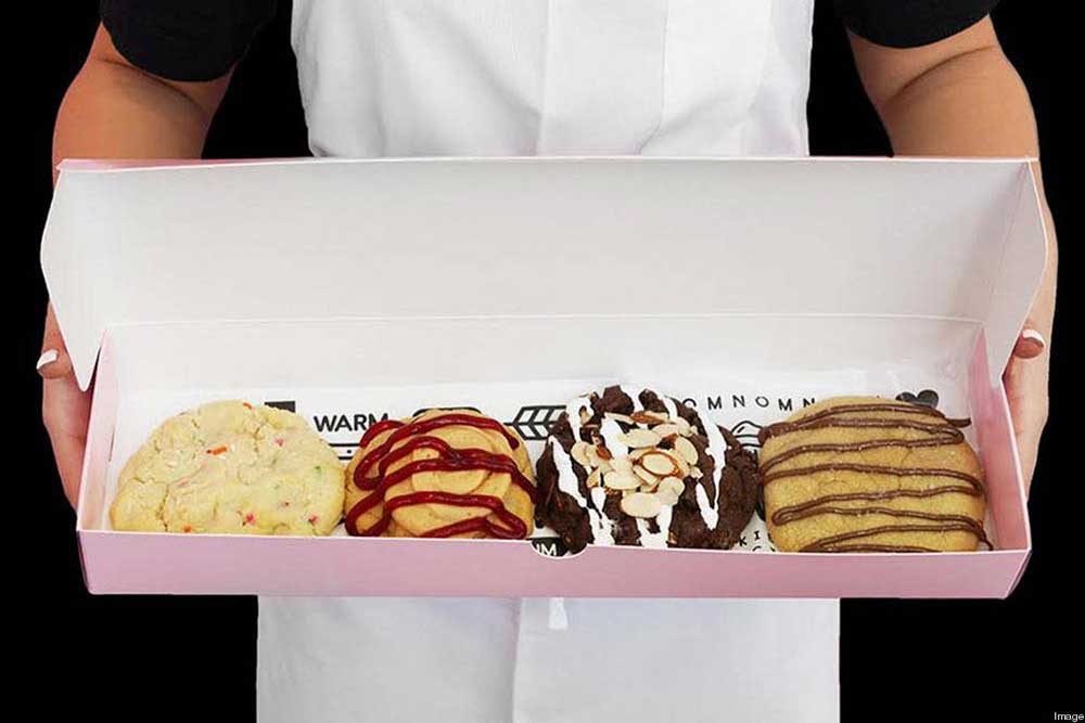 An employee holding a pink box of four cookies with various toppings 