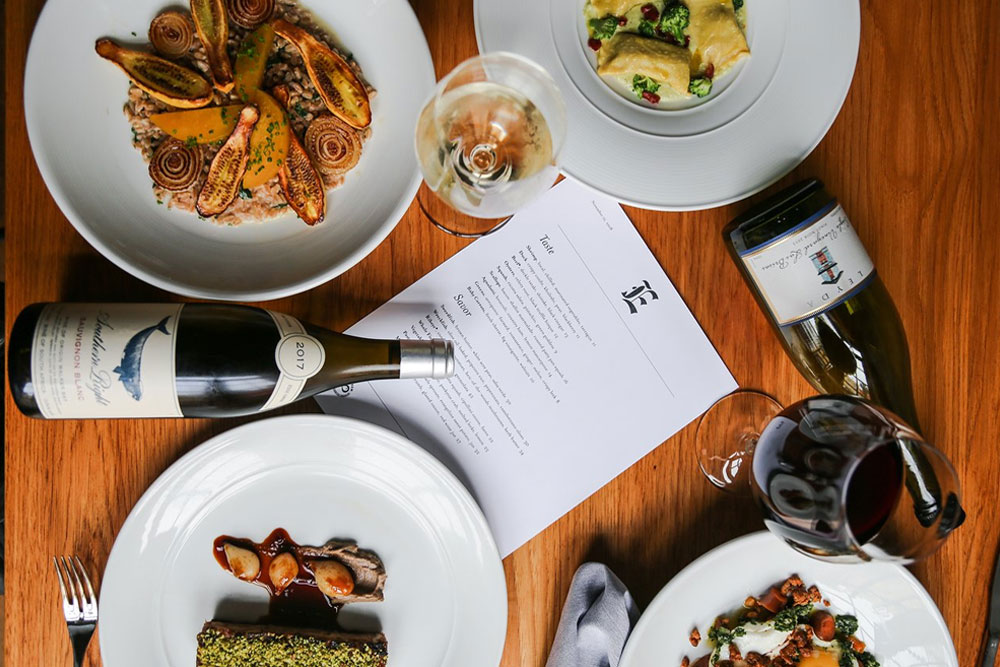 An overhead shot of several dishes, bottles and glasses of wine, and a menu