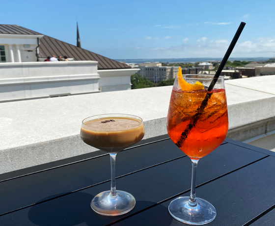 An espresso martini and an aperol spritz with a straw on a table overlooking the Charleston skyline