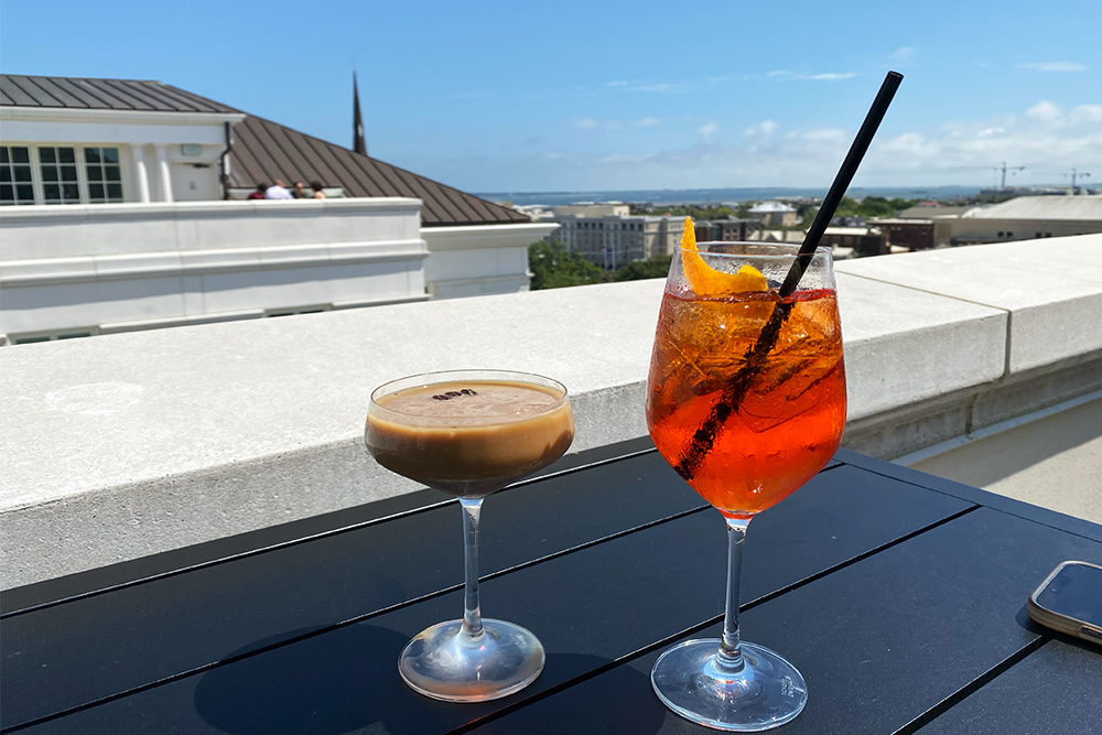 An espresso martini and an aperol spritz with a straw on a table overlooking the Charleston skyline