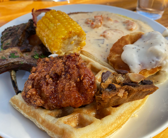 Plate full of breakfast foods, including lamb chops, waffles, biscuits, fried chicken, corn, and more