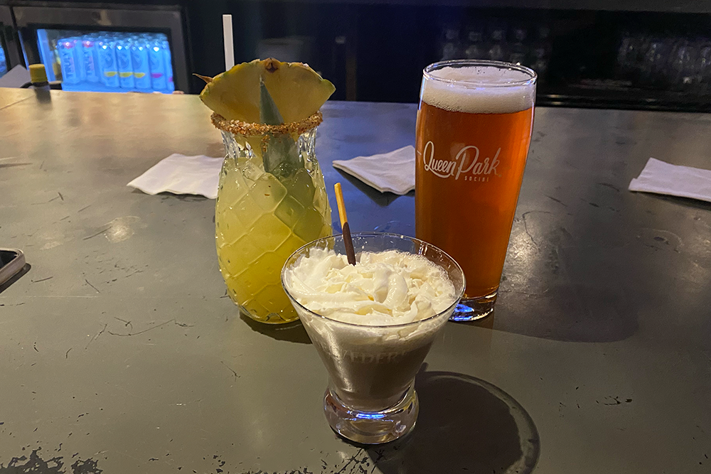 Three drinks on a bar: one with a pineapple wedge, one with whipped cream on top and a candy hanging out, and a beer