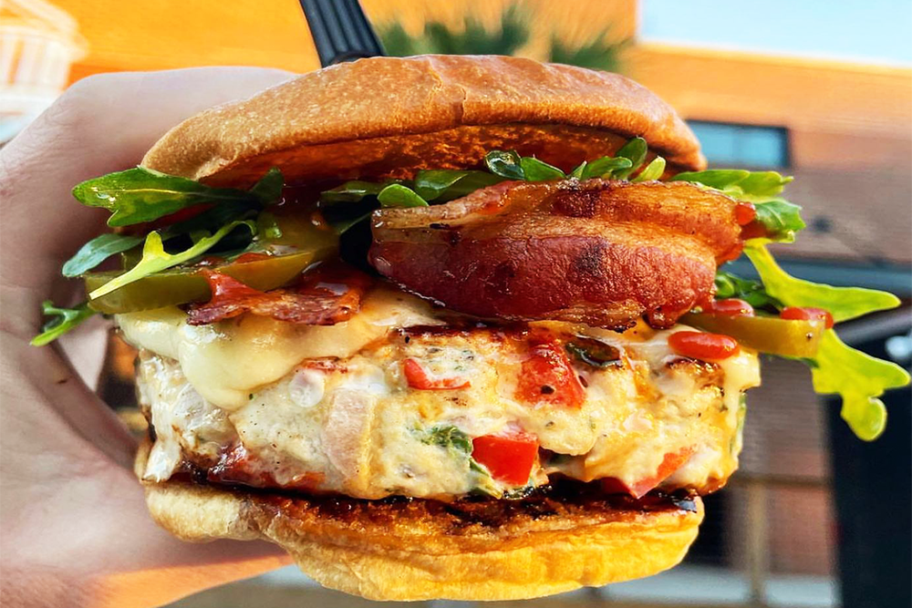 A close-up of a hand holding a turkey burger topped with cheese, peppers, bacon, and lettuce