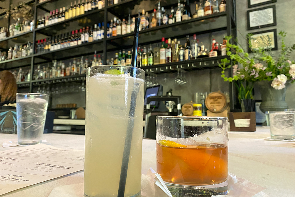 Two drinks on a bar with several shelves of liquor in background