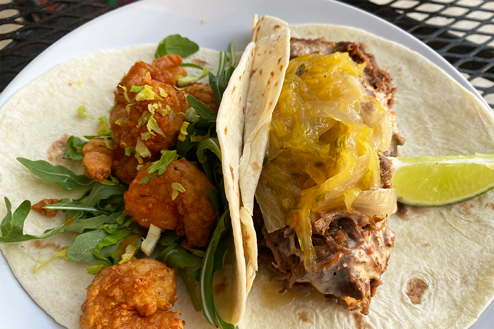 Two tacos, one with breaded shrimp and lettuce and the other with short rib and caramelized onion with a lime wedge