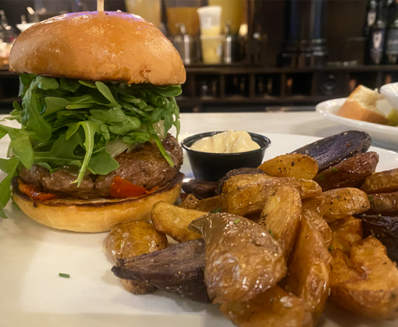 Burger with arugula, roasted tomato, gruyere cheese, and more with a side of fingerling potatoes and an aioli in a black cup