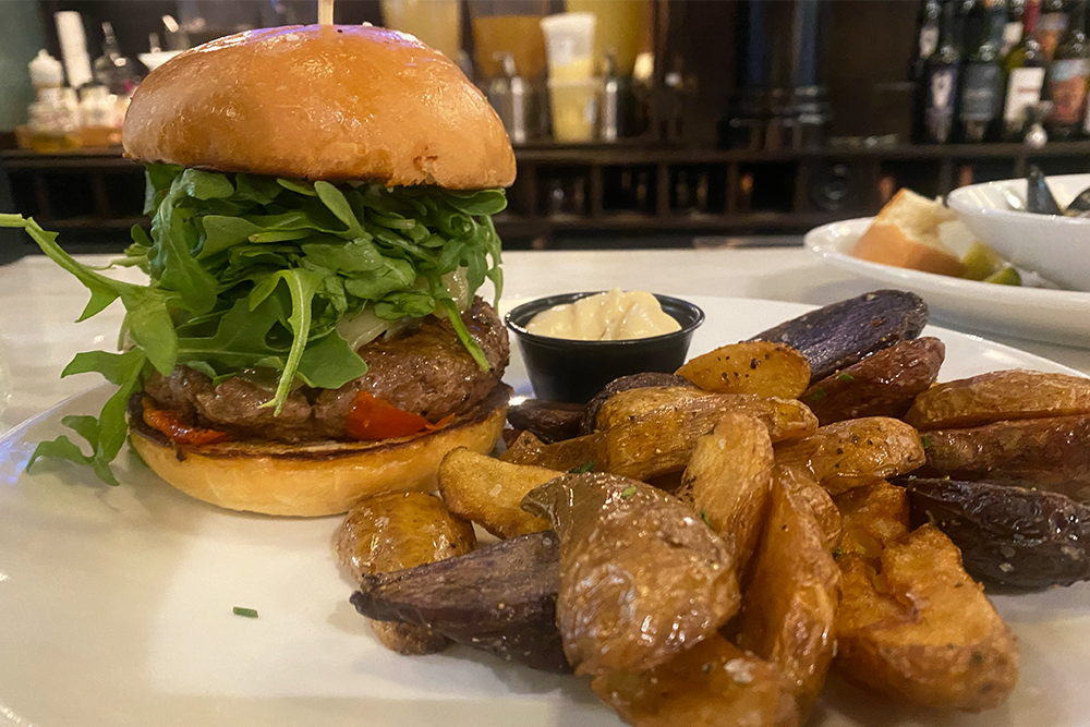 Burger with arugula, roasted tomato, gruyere cheese, and more with a side of fingerling potatoes and an aioli in a black cup
