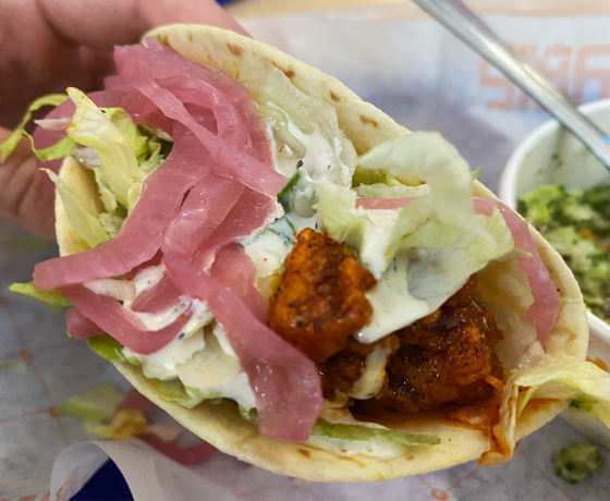 Hand holding a soft hot chicken taco, topped with shredded lettuce, pickled onions, and ranch