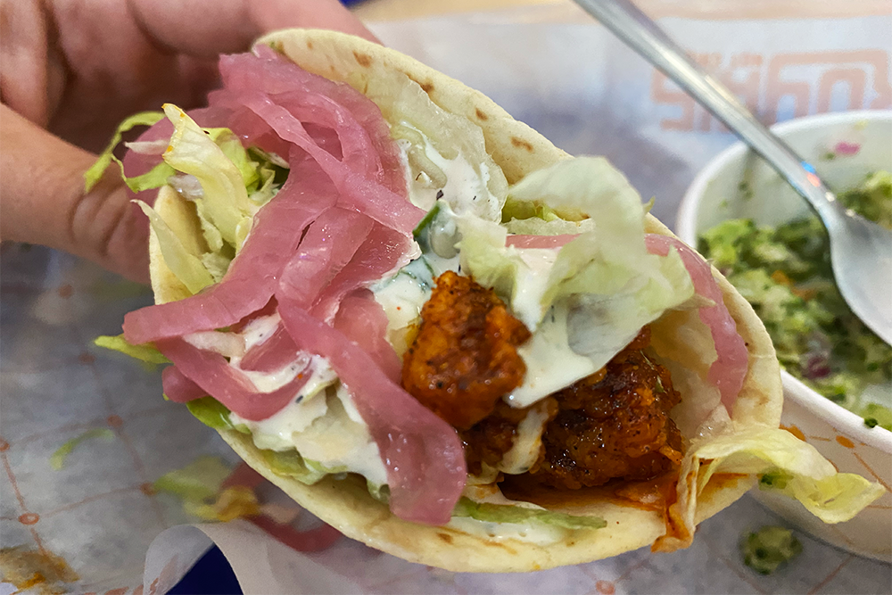 Hand holding a soft hot chicken taco, topped with shredded lettuce, pickled onions, and ranch
