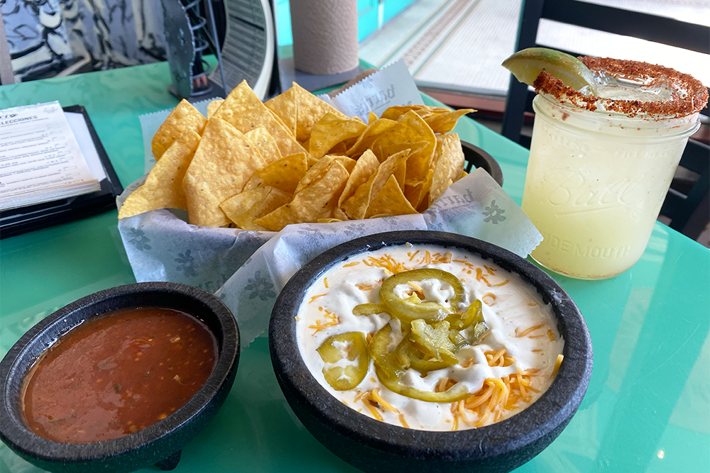 Basket of chips with a bowl of salsa and queso with peppers. There is a glass of margarita behind them