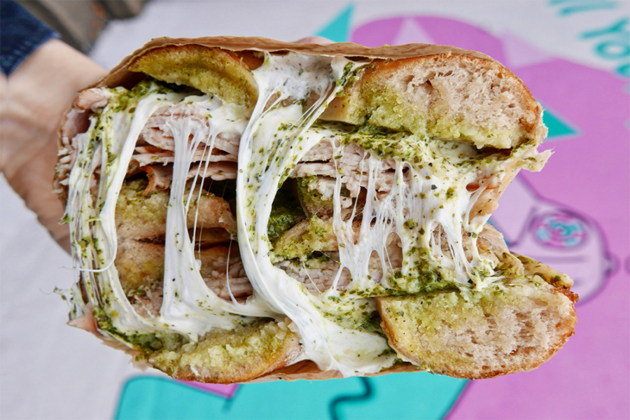 A bagel sandwich sliced in half, with melty cheese, turkey, red pepper relish, and pesto against a pink and blue background